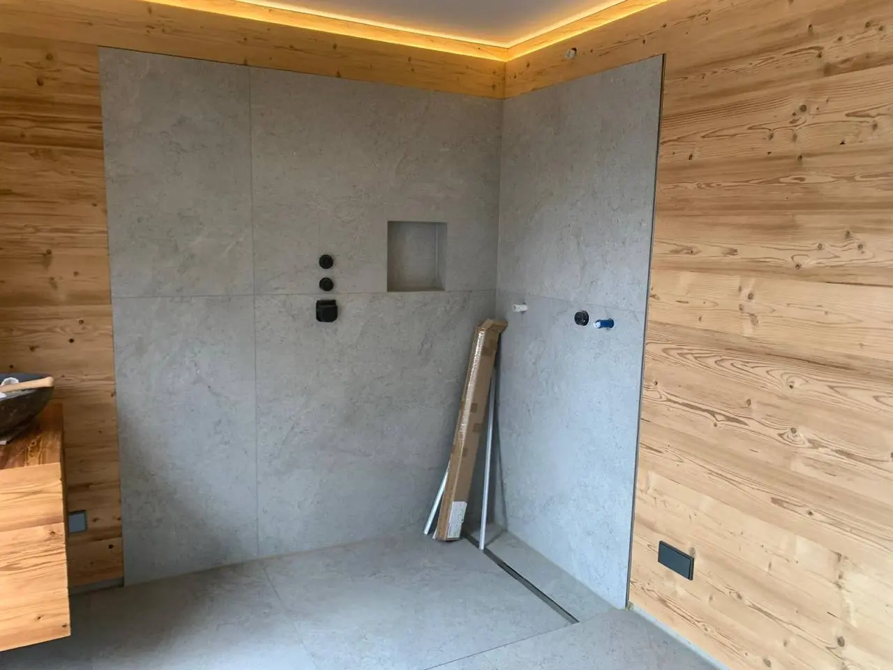 Duschbereich mit großformatigen grauen Platten, Wandnische, umlaufender Holzverkleidung und indirektem Licht