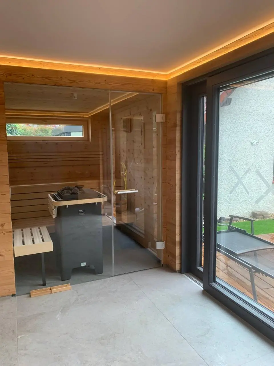 Private Holzsauna mit Glasfront, Sitzbank, Saunaofen und umlaufender indirekter Deckenbeleuchtung